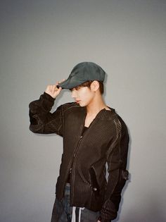 a young man wearing a black jacket and hat standing in front of a gray wall