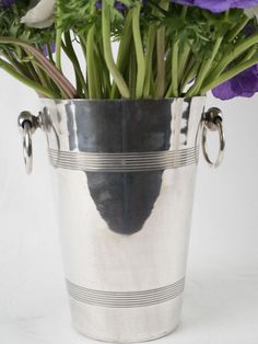 purple flowers are in a silver bucket on a white background with two handles and one flower is hanging from the side