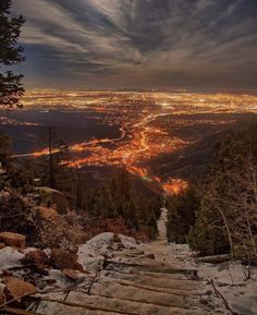 a wooden walkway leading up to the top of a snowy hill at night with lights in the distance