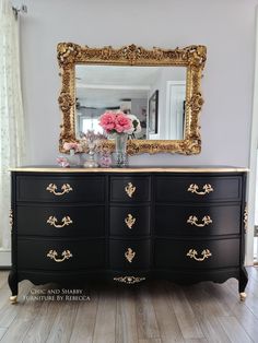 a black and gold dresser with pink flowers on top in front of a large mirror