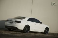 a white car is parked in front of a wall and has black rims on it