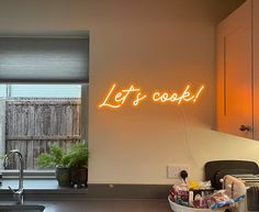 a neon sign that says let's cook on the wall above a kitchen sink