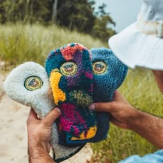 a person holding three knitted animal masks in front of grass and dirt area with trees in the background