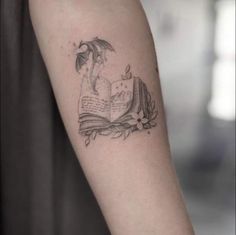 an open book tattoo on the arm with a dragon flying over it, surrounded by flowers and leaves