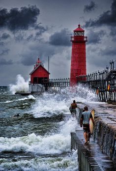 two surfers walk into the ocean near a red light house with waves crashing against it