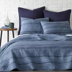 a bed with blue comforters and pillows in front of a white brick wall,