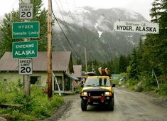 a yellow truck driving down a dirt road next to a forest covered mountain with snow capped mountains in the background