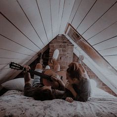 two people sitting on a bed playing the guitar in their attic bedroom, while another person is laying down next to them