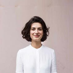 a woman standing in front of a wall wearing a white shirt and black pants with her hands in her pockets