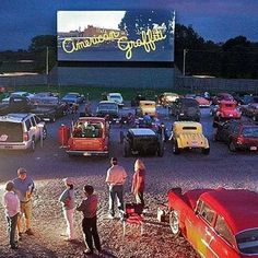 a group of people standing in front of a movie screen with cars parked next to it