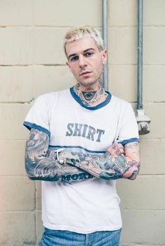 a man with tattoos standing in front of a brick wall and holding his arms crossed