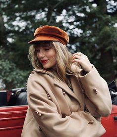 a woman sitting on the back of a red car wearing a tan coat and hat