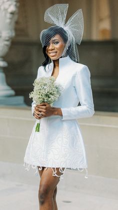 a woman in a white dress and hat holding a flower bouquet while walking down the street