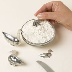 a person is spooning flour into a bowl with two ducks on the side and three silver spoons next to it