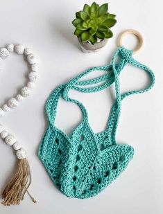 a crocheted bag next to a plant and beaded necklace on a white surface