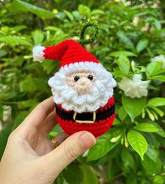 a hand holding a small red and white santa clause ornament in front of some bushes