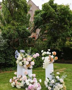 two tall white vases with flowers are on the grass in front of some trees