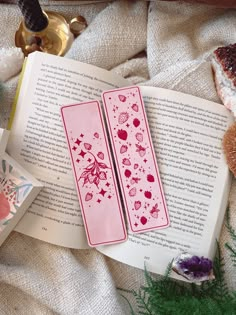 two pink bookmarks sitting on top of an open book next to some bread and flowers