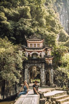 two people sitting on the edge of a small bridge in front of a mountain stream