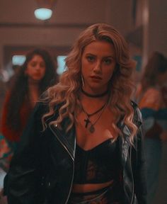 a woman with blonde hair wearing a black top and leather jacket standing in front of other women