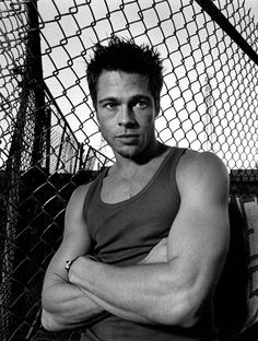 a black and white photo of a man leaning against a fence with his arms crossed