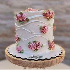 a white cake with pink roses on it sitting on top of a wooden stand next to a brick wall