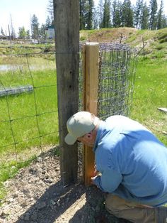 a man in blue shirt and white hat leaning against fence with his hands on the post