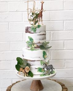 a three tiered cake decorated with animals and greenery on a wooden stand next to a brick wall