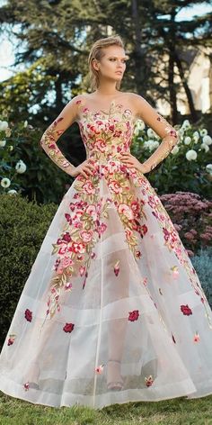 a woman in a long dress with flowers on it is standing near some bushes and trees