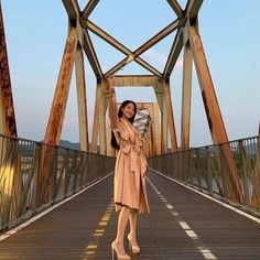 a woman is standing on a bridge with her arms in the air and she is wearing a tan coat