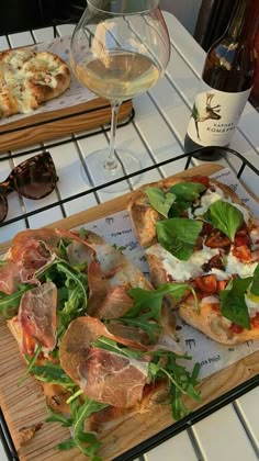 two pizzas with greens and cheese on a cutting board next to glasses of wine