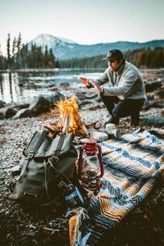 a man sitting on top of a blanket next to a campfire near a lake