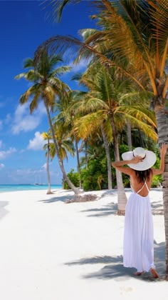 a woman in a white dress and straw hat standing on the beach with palm trees