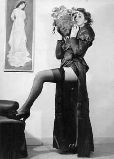 an old photo of a woman sitting on a chair with her legs crossed and holding flowers