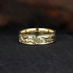 a gold wedding band with green stones on the side and leaves in the middle, sitting on a black surface