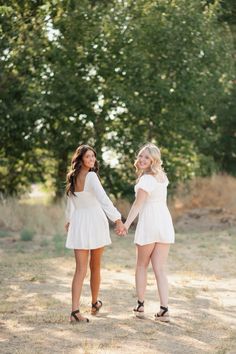 two women in white dresses holding hands and smiling at each other with trees in the background