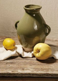 a green vase sitting on top of a wooden table next to two lemons and an apple