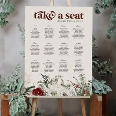 an easel with flowers and greenery on it is next to a sign that says take a seat