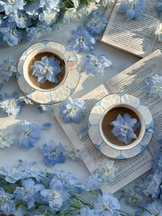 two cups of coffee are sitting next to an open book with blue flowers on it