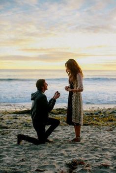 a man kneeling down next to a woman on top of a sandy beach near the ocean