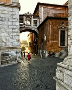 two people walking down an old cobblestone street under a bridge in the city