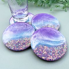 three purple and blue coasters with glitter on them next to a glass vase filled with greenery