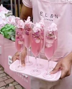 a person holding a tray with five glasses filled with pink liquid and flowers on it