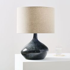 a black table lamp with a white shade on it and a glass vase next to it