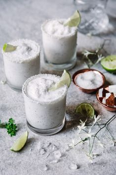 three glasses filled with margarita cocktails next to lime wedges and small wooden spoons