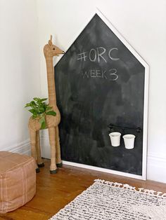 a giraffe standing next to a blackboard with writing on it and potted plants