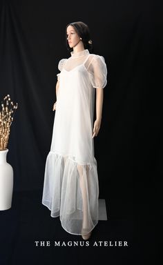 This elegantly crafted dress features a delicate organza overlay that adds a whimsical touch to any occasion. The translucent organza top layer boasts puffed sleeves and a high neckline, giving a vintage-inspired yet modern silhouette. Underneath, a sleek, simple Shenton inner slip ensures coverage while maintaining a graceful, lightweight look.  Key Features: - Outer Layer: Lightweight organza fabric for a sheer, ethereal appearance - Sleeves: Short puff sleeves for added volume and a soft roma Sheer Organza Mesh Evening Dress, White Organza Tulle Fabric With Ruffles, Elegant Sheer Mesh Dress In Organza, Elegant Organza Dresses With Overlay, White Sheer Sleeves Maxi Dress For Summer, White Fitted Organza Maxi Dress, White Sheer Sleeves Summer Maxi Dress, Summer Organza Dress With Overlay, White Maxi Dress With Sheer Sleeves For Summer