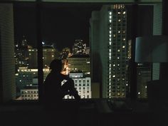 a man sitting in front of a window looking out on the city at night time