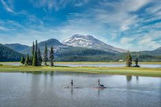 two people stand on surfboards in the water near mountains and trees, while another person stands on a paddle board