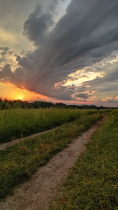 the sun is setting over a grassy field with a dirt path in front of it